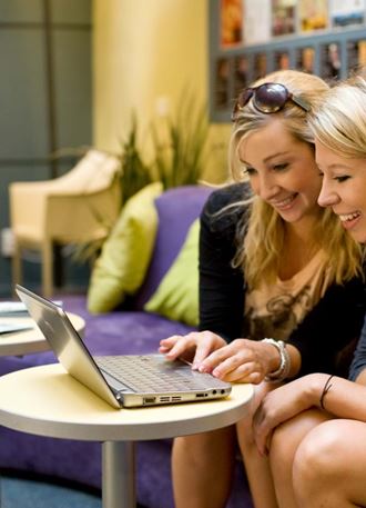 Two young women at YHA Wellington smile and check their laptop together in the shared hostel dining area. 